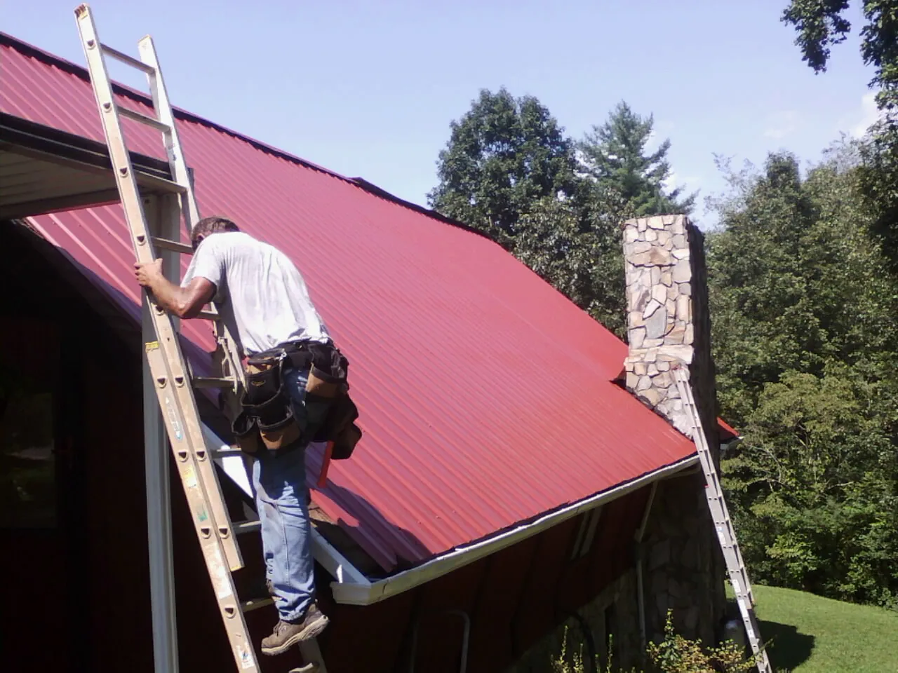 Roofer on a ladder beside a red metal roof and stone chimney — Soffit & Fascia Repair by Roofing Brothers in Farmersburg
